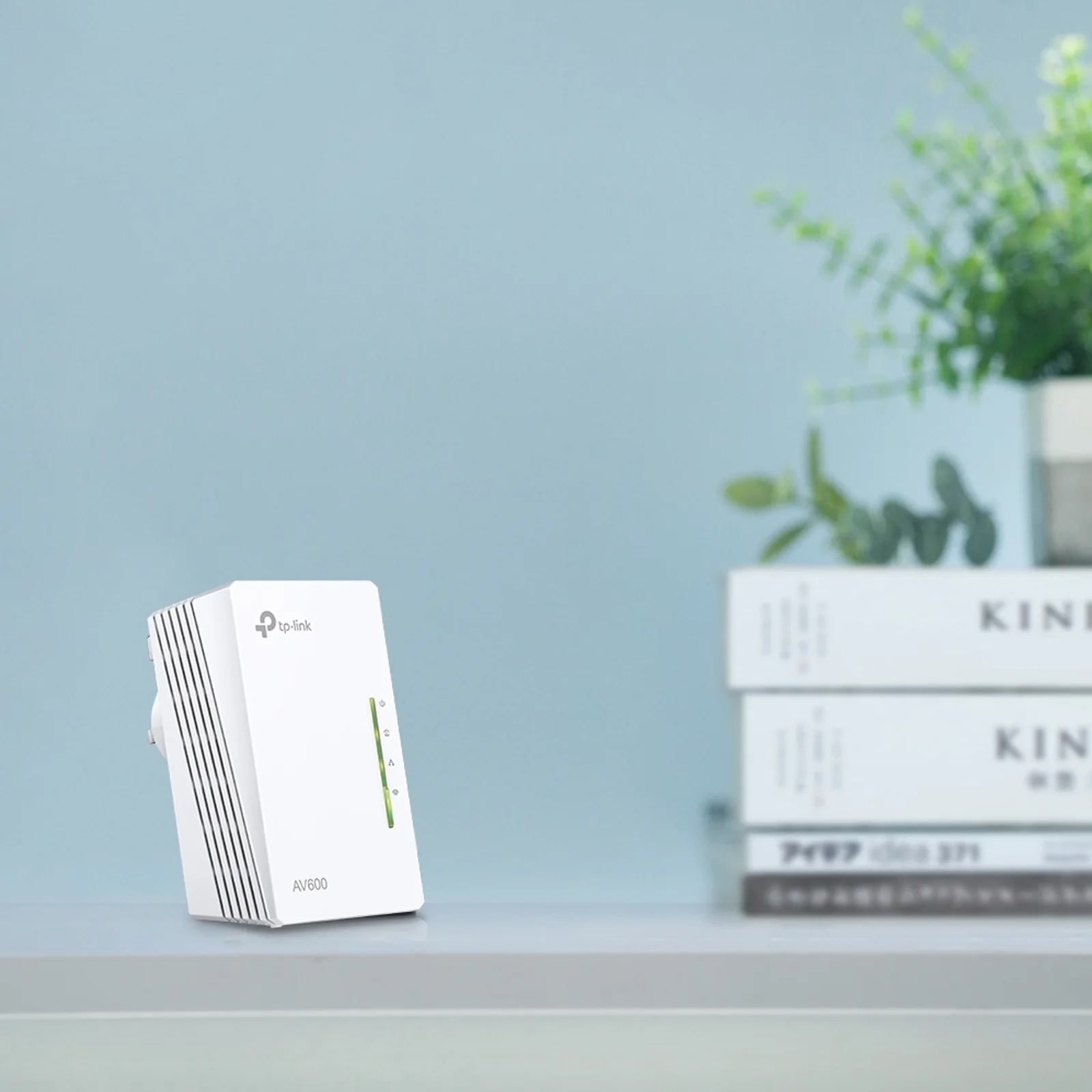 Adaptateur TP-Link TL-WPA4220 AV600 posé sur une étagère avec des livres empilés en arrière-plan, fond bleu gris, mise en situation bureau