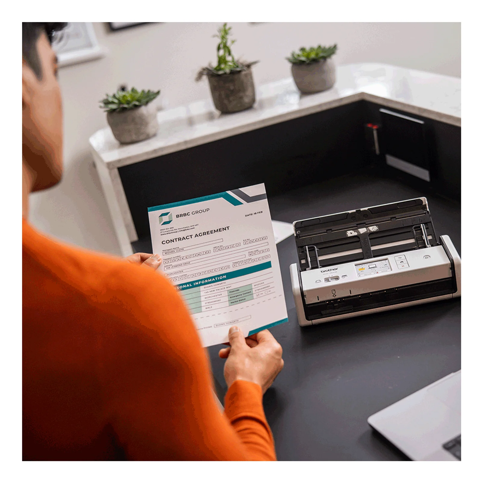 Personne en pull orange tenant un contrat près d'un scanner ADS-1800W posé sur bureau noir avec plantes en arrière-plan