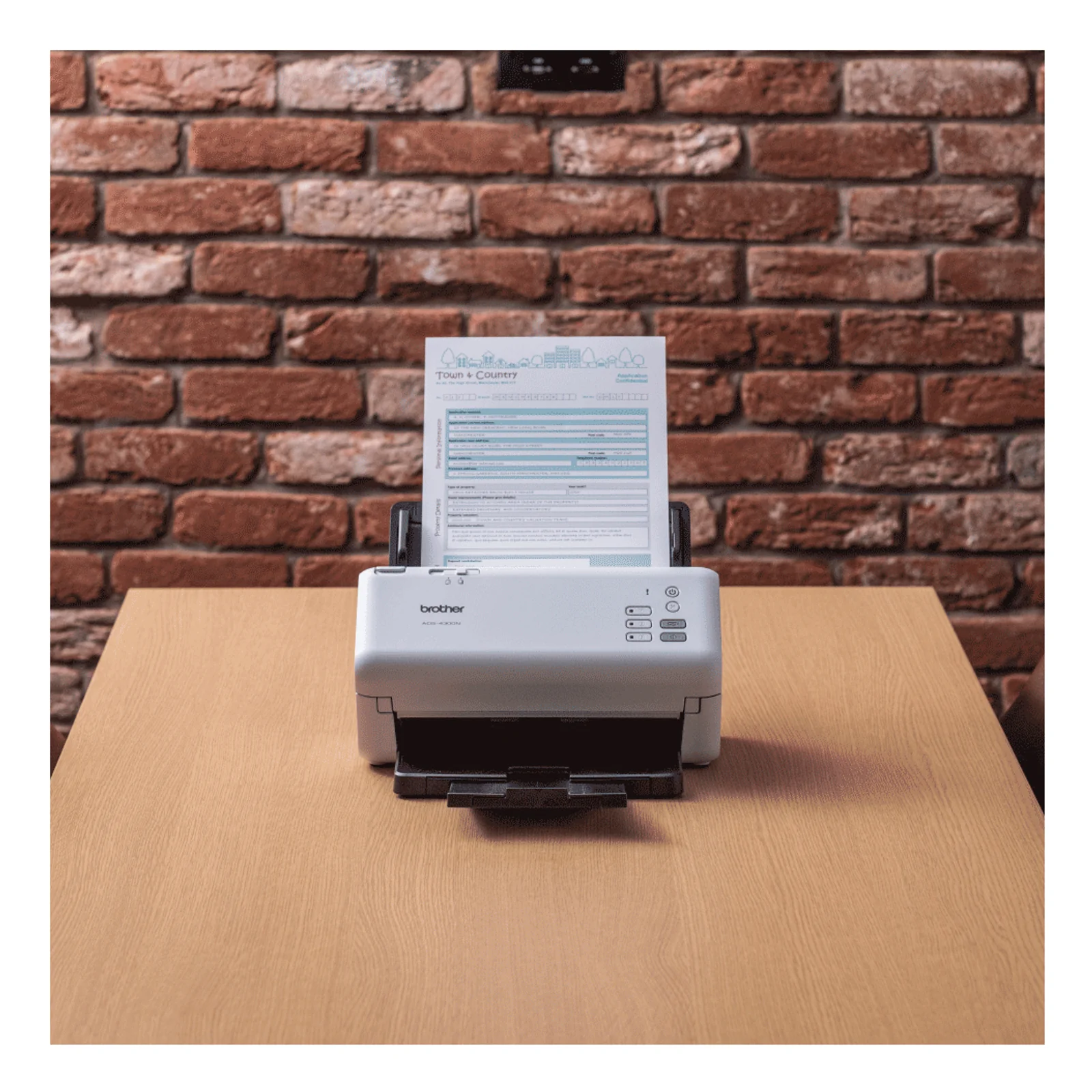 Scanner Brother ADS-4300N posé sur une table en bois clair devant un mur de briques rouges, formulaire en cours de scan