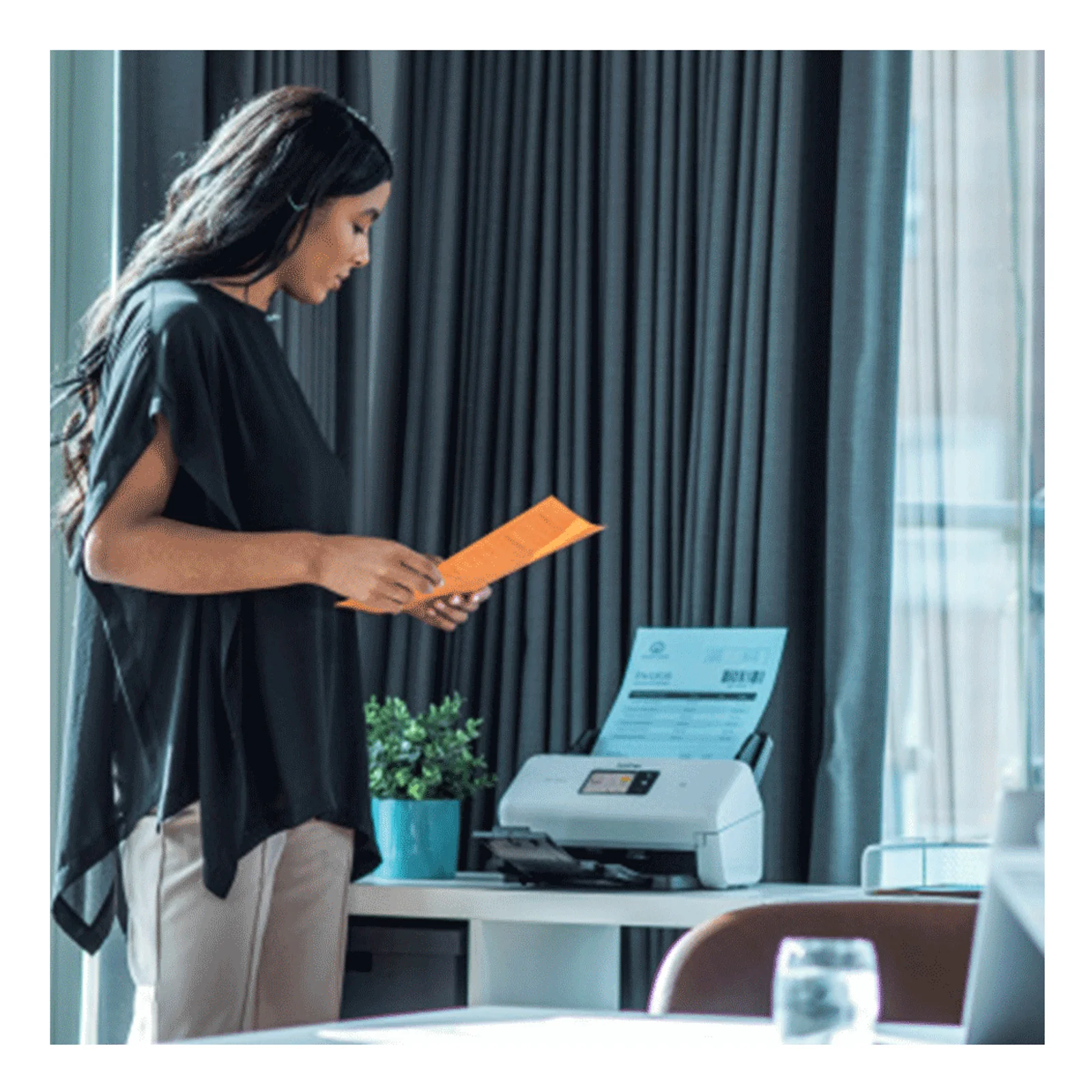 Femme en haut noir tenant une enveloppe orange devant un scanner Brother ADS-4550W posé sur un bureau de bureau professionnel