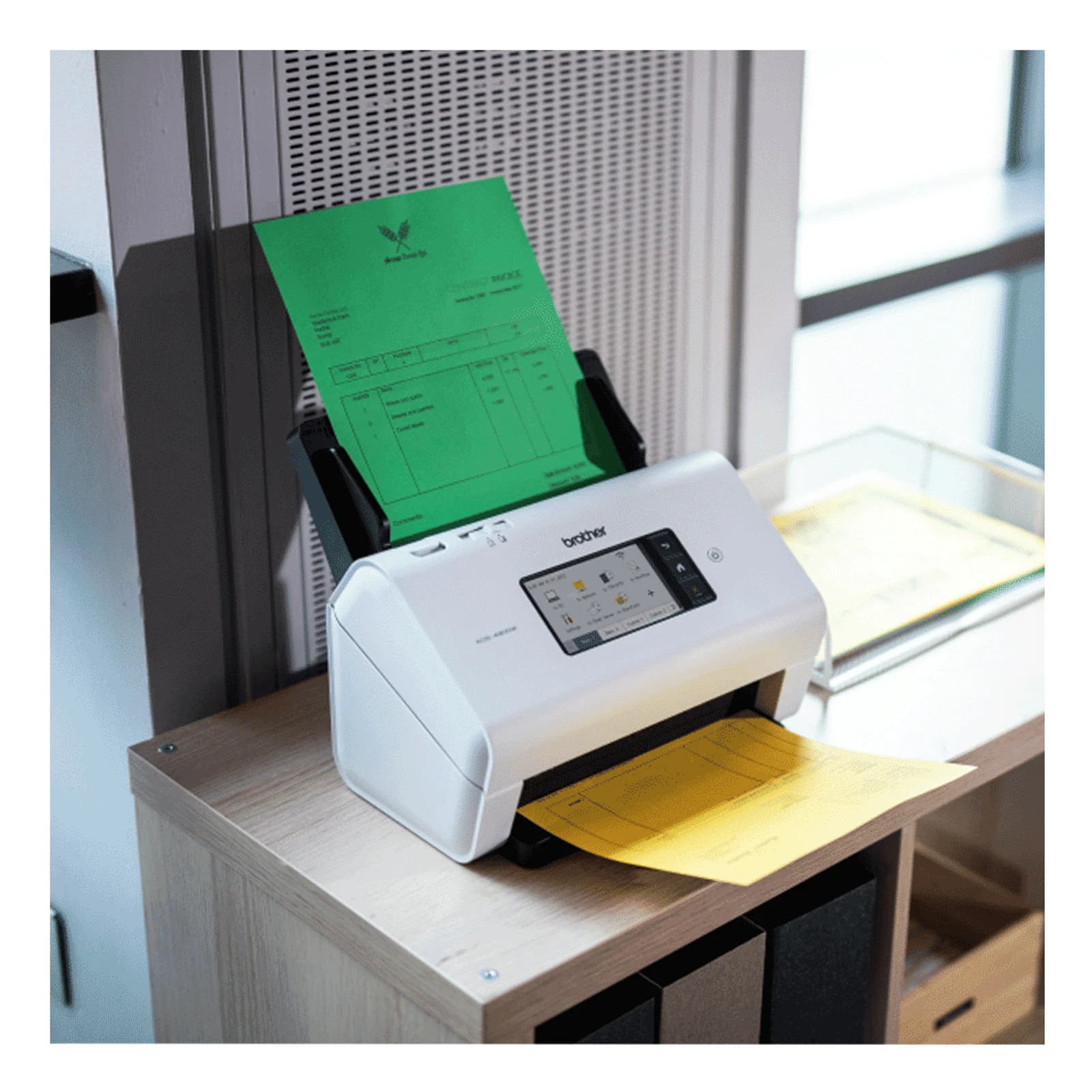 Scanner Brother ADS-4900W posé sur un meuble de bureau avec document vert en alimentation et feuilles jaunes en sortie sur le côté