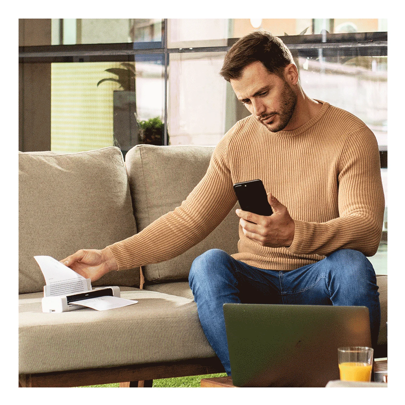Homme assis dans un canapé tenant un smartphone et chargeant une feuille dans le scanner Brother DS-940DW posé sur la table