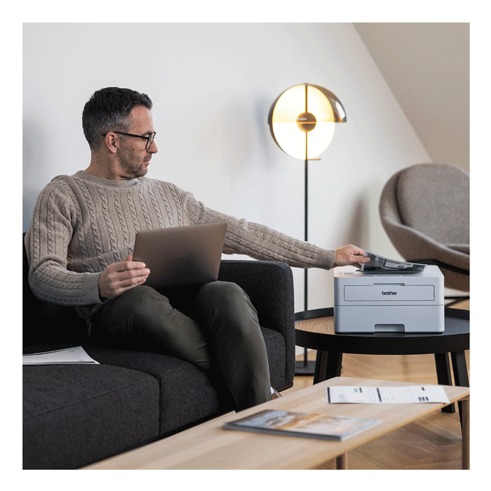 Brother HL-L2865DW en mise en situation salon avec utilisateur sur canape lancant une impression depuis son ordinateur portable