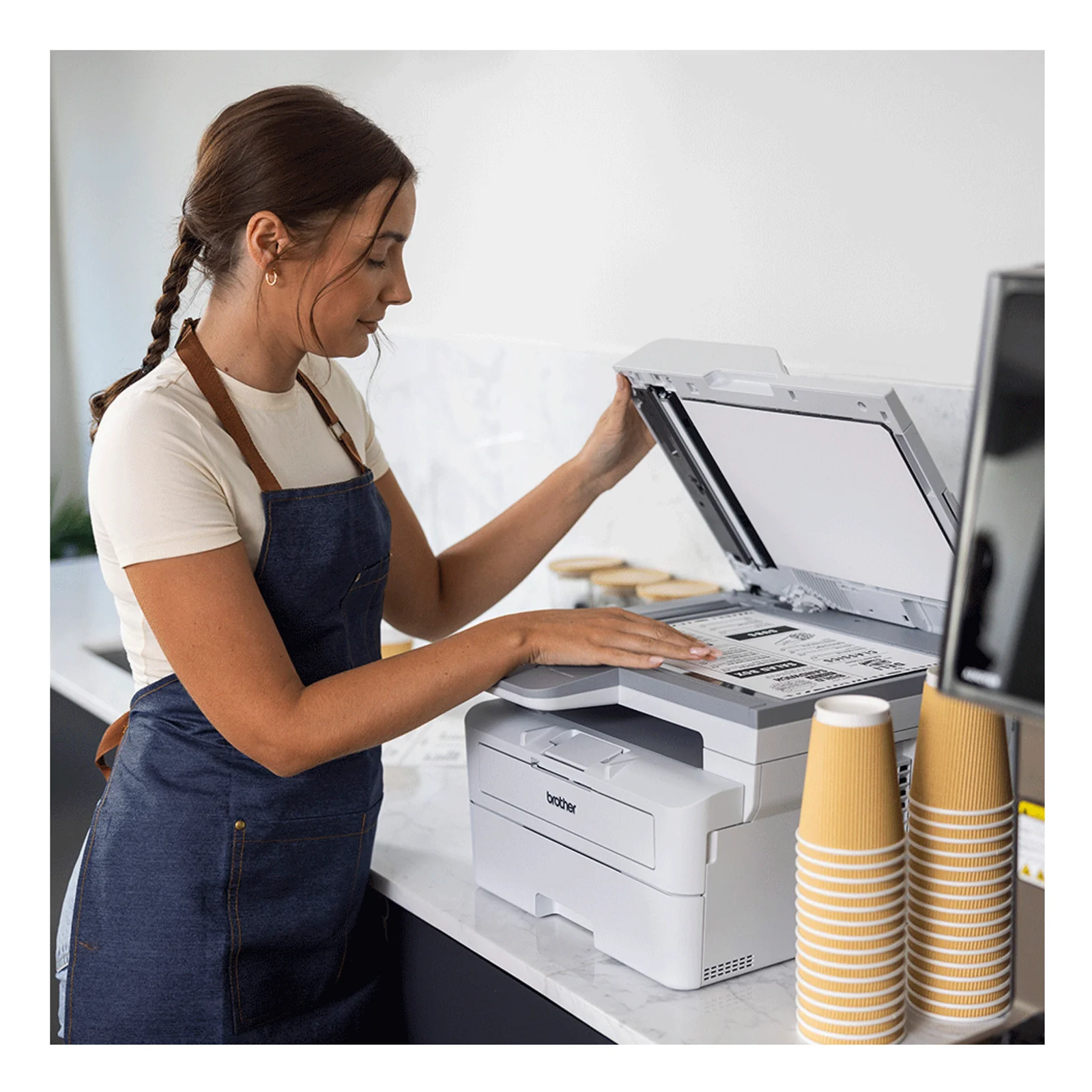 Femme en tablier bleue soulevant le capot du scanner du MFC-L2960DW dans un comptoir avec gobelets jetables empilés