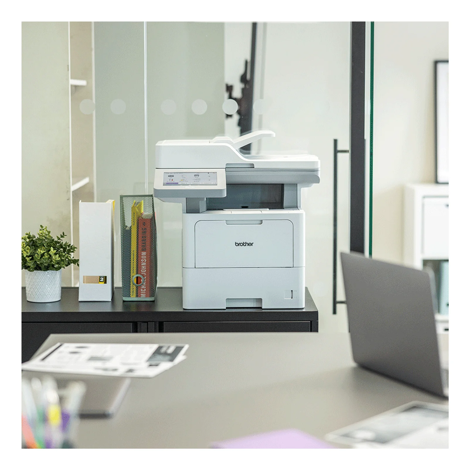 Multifonction Brother MFC-L6910DN posé sur un meuble de bureau clair avec ordinateur portable et plante en arrière-plan