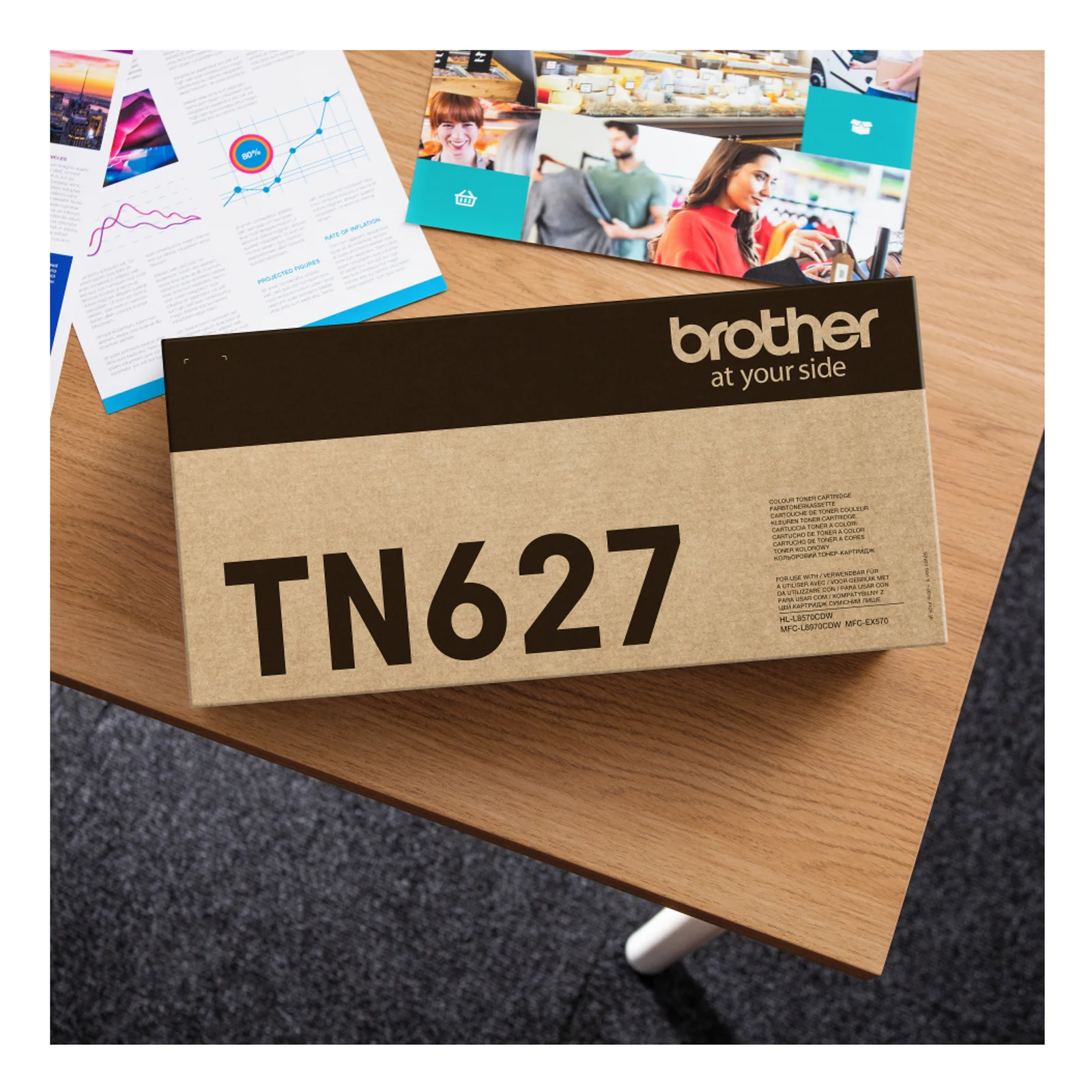 Boîte du toner Brother TN627 posée sur un bureau bois à côté de plaquettes commerciales colorées