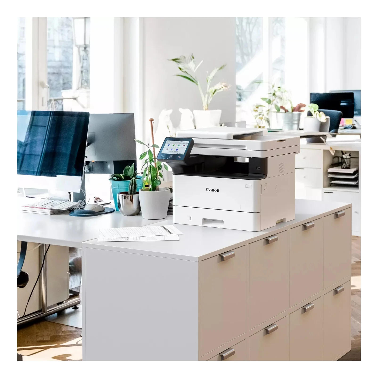 Canon i-SENSYS MF463dw II posé sur un meuble dans un bureau lumineux avec écrans et plantes vertes