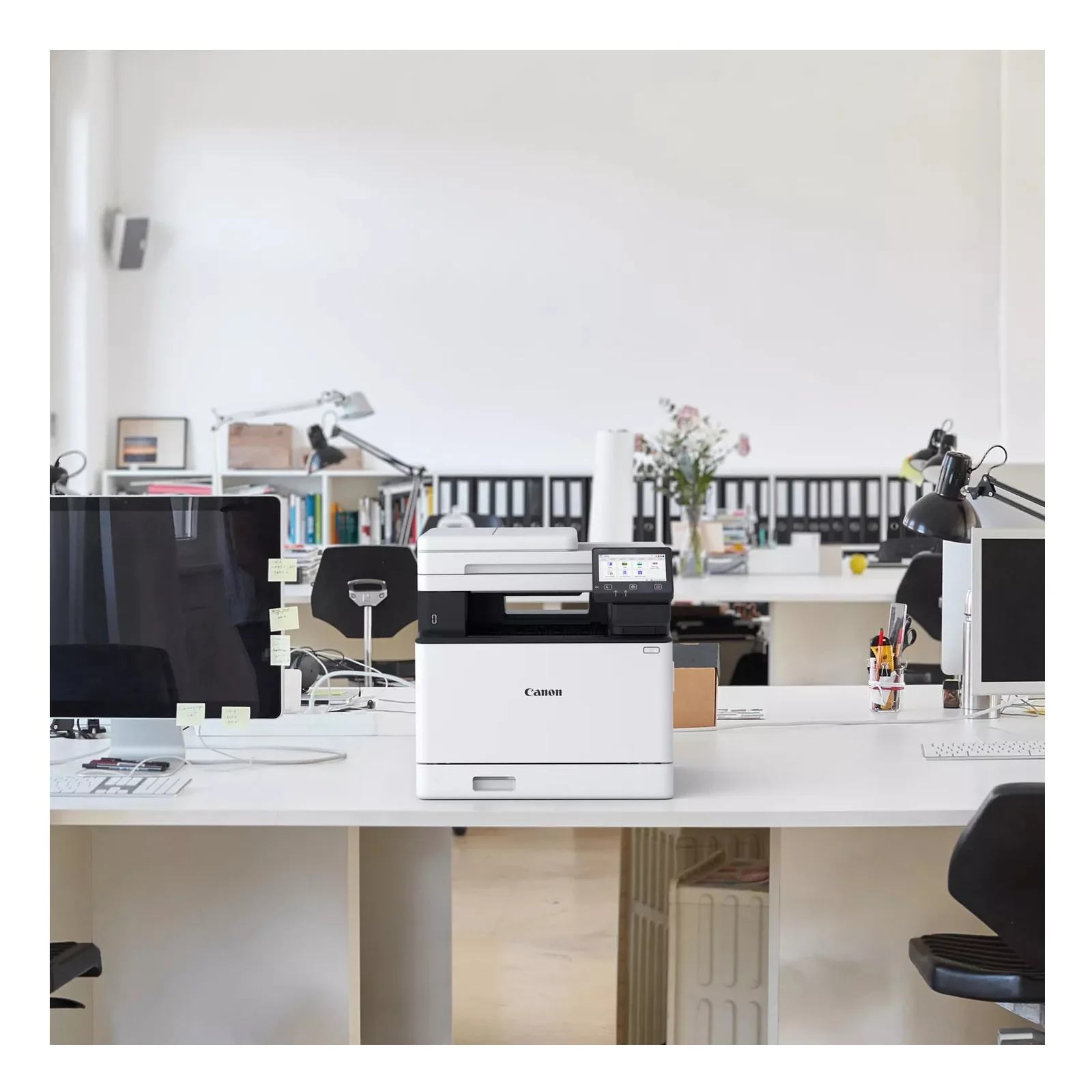 Canon MF754Cdw II posé sur un bureau partagé en open space avec deux écrans, lampes articulées et plante en arrière-plan