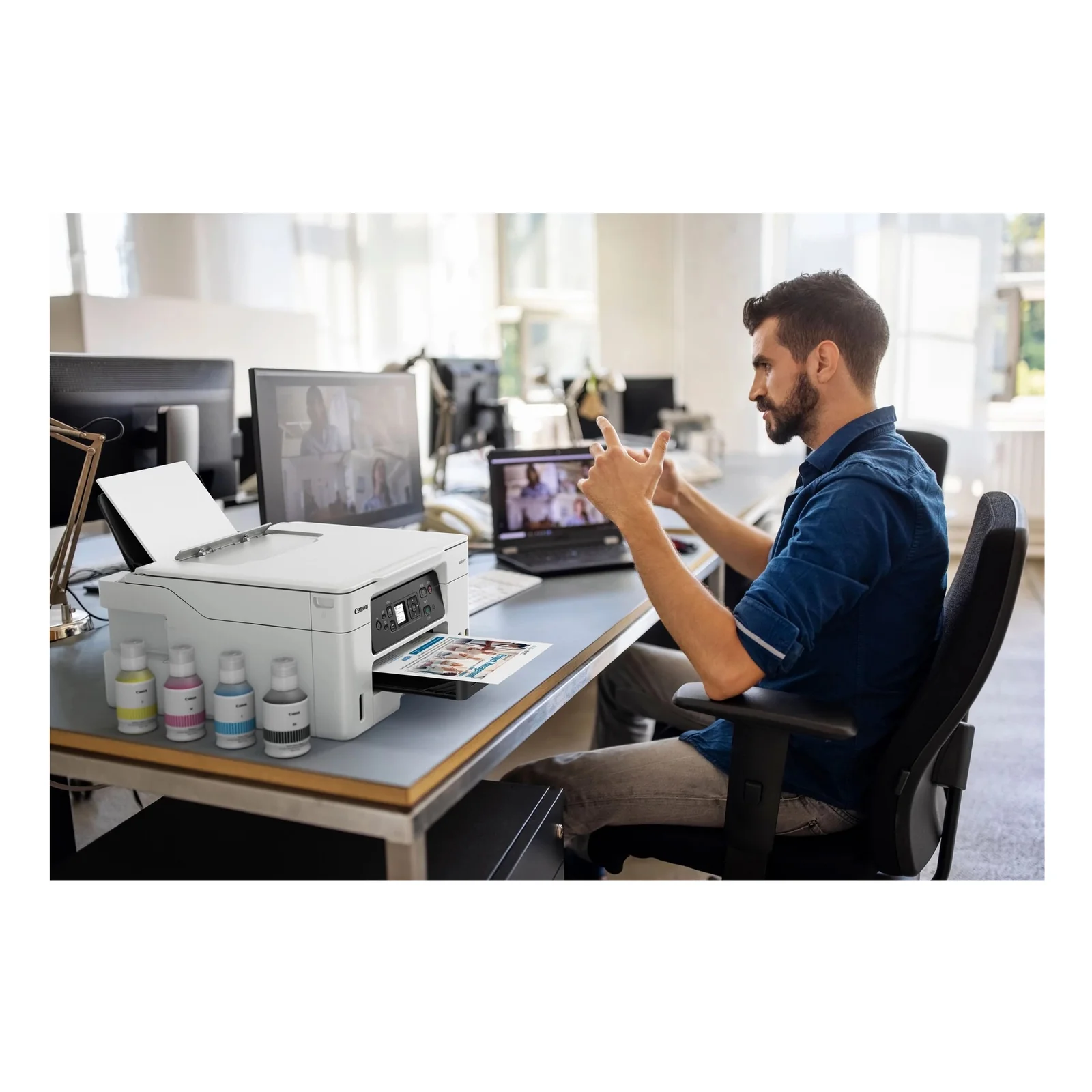 Homme en chemise bleue en réunion vidéo avec la MAXIFY GX3050 et bouteilles d'encre sur le bureau