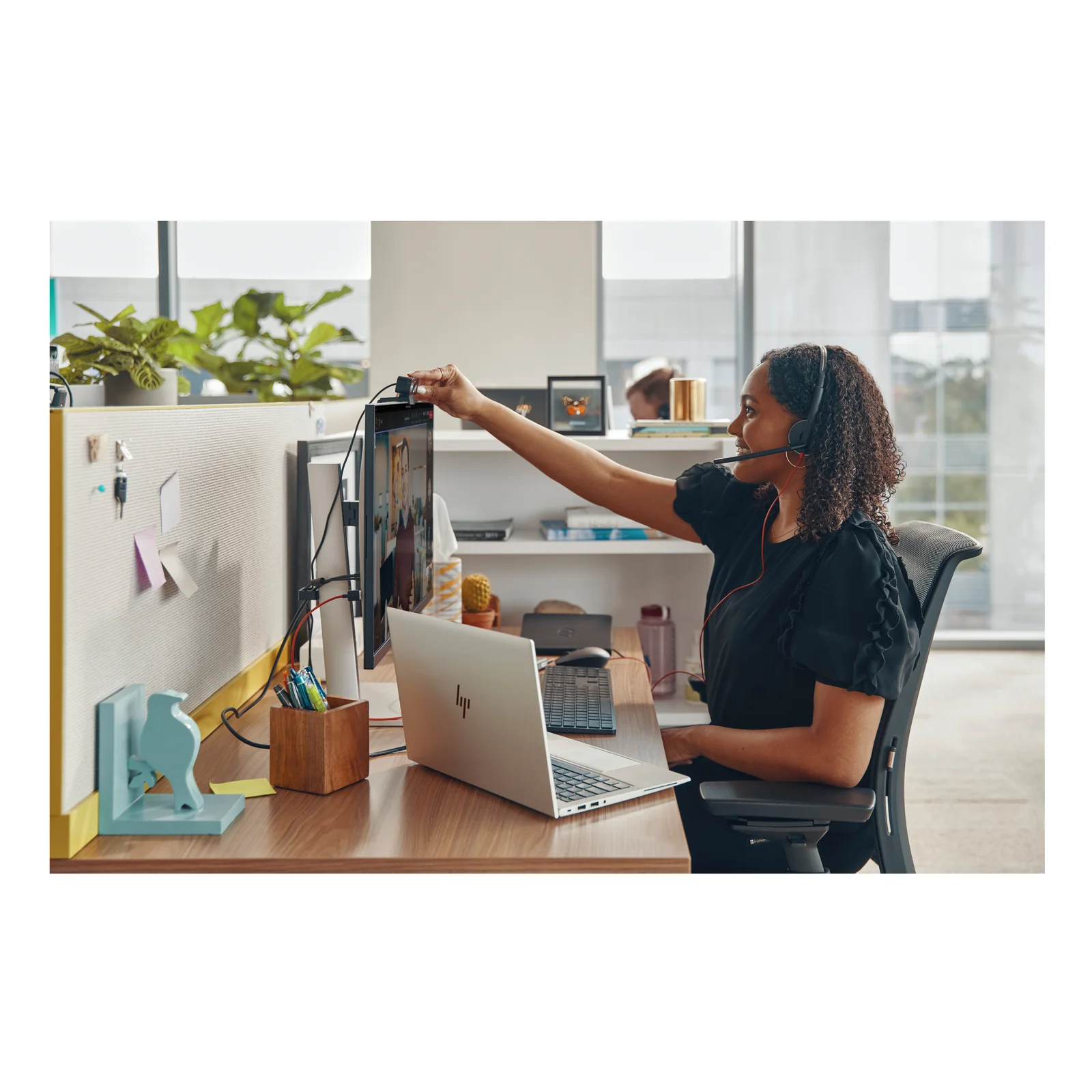 Femme assise à son bureau portant le HP Poly Blackwire 3215 monaural, ajustant un moniteur lors d'un appel, ordinateur portable HP et environnement de bureau open space