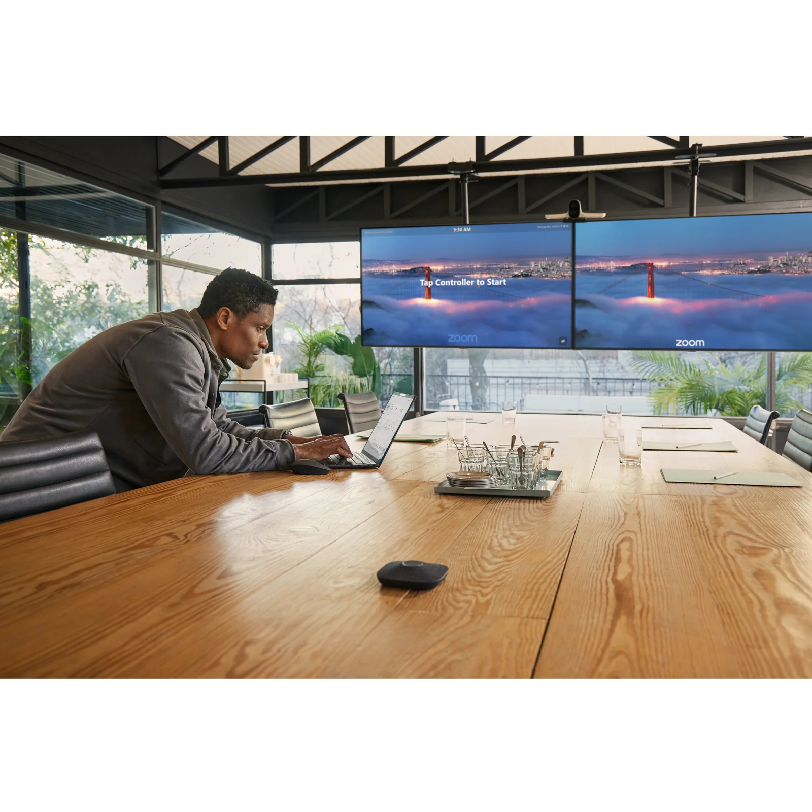 Vue d'ensemble d'une grande salle de conférence avec un homme travaillant sur ordinateur portable, deux écrans Zoom en arrière-plan, pont audio HP Poly visible sur la table.