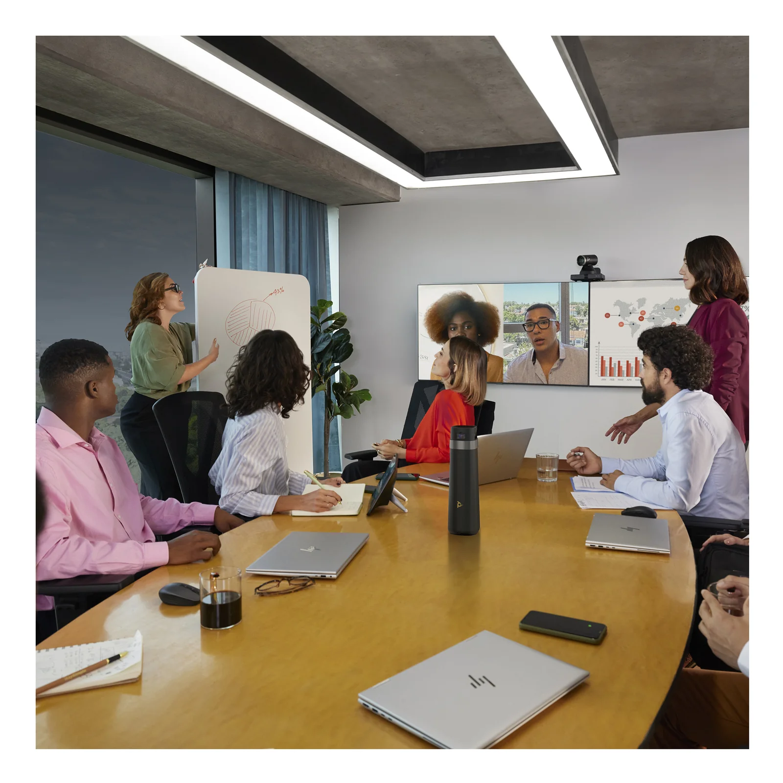 Photo de salle de réunion avec présentateur au tableau blanc, équipe assise et participants en visioconférence sur écran mural, caméra Poly visible en hauteur