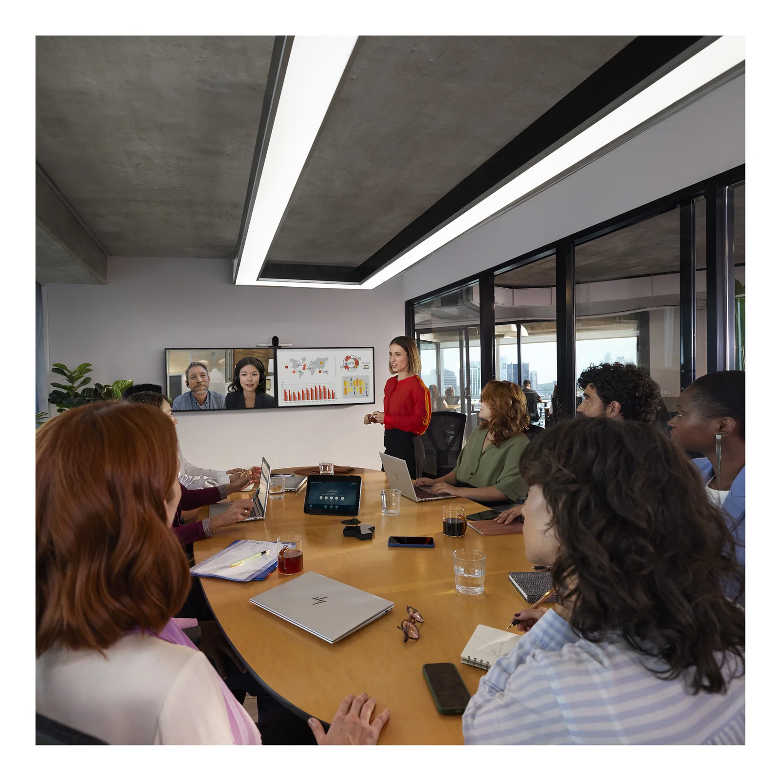 Grande salle de réunion avec HP Poly Studio G62, groupe de collaborateurs autour d'une table ovale avec tablette de contrôle, écran mural affichant des participants distants