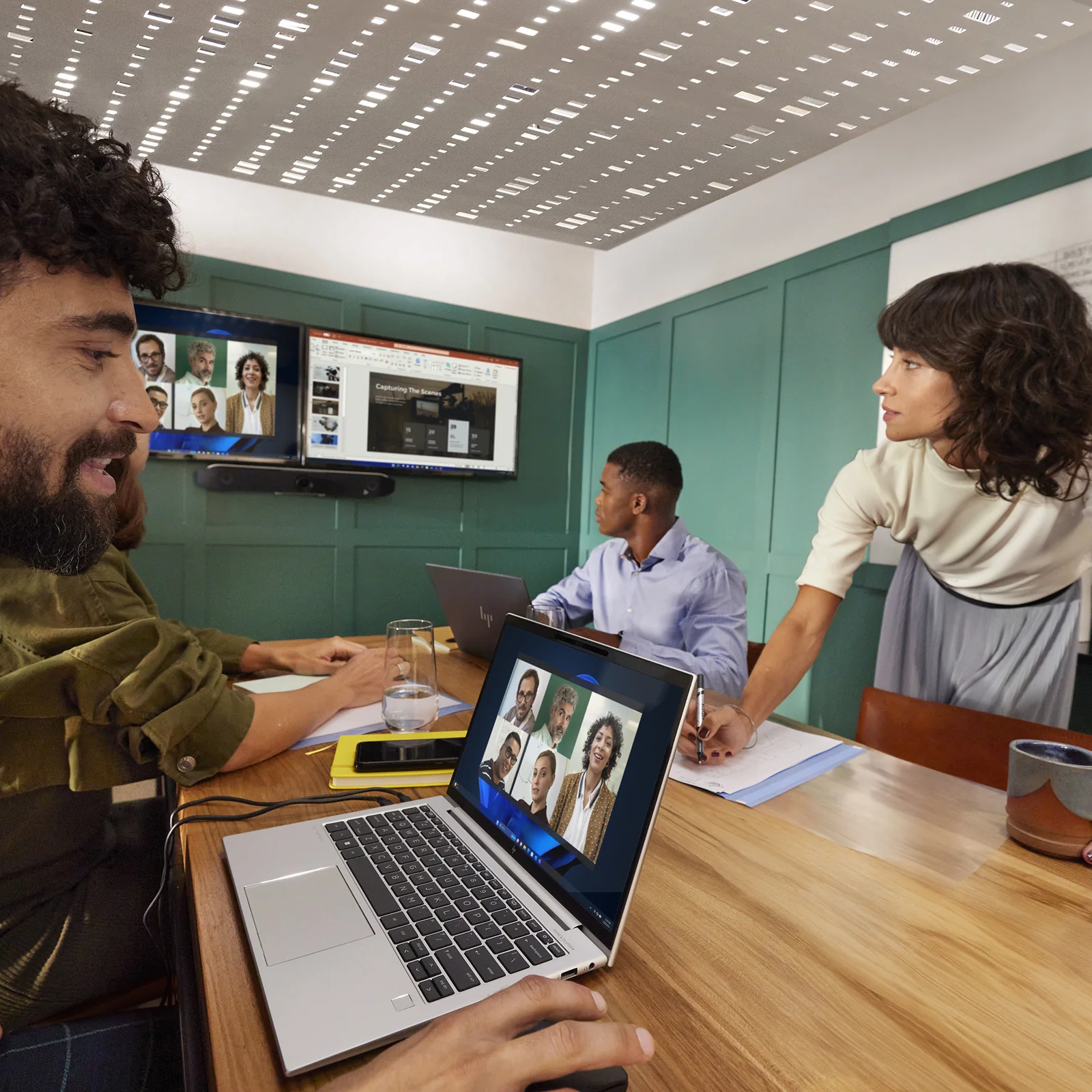 Grande salle de réunion avec HP Poly Studio V52 sur écran mural, groupe de participants autour d'une table en réunion hybride avec appel vidéo Teams