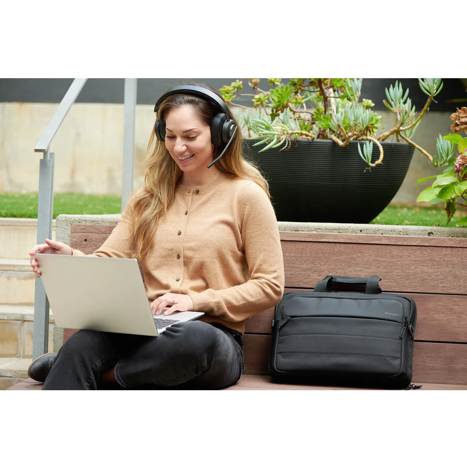Femme assise en extérieur avec un casque audio, ordinateur portable sur les genoux, sacoche Kensington EQ noire à côté