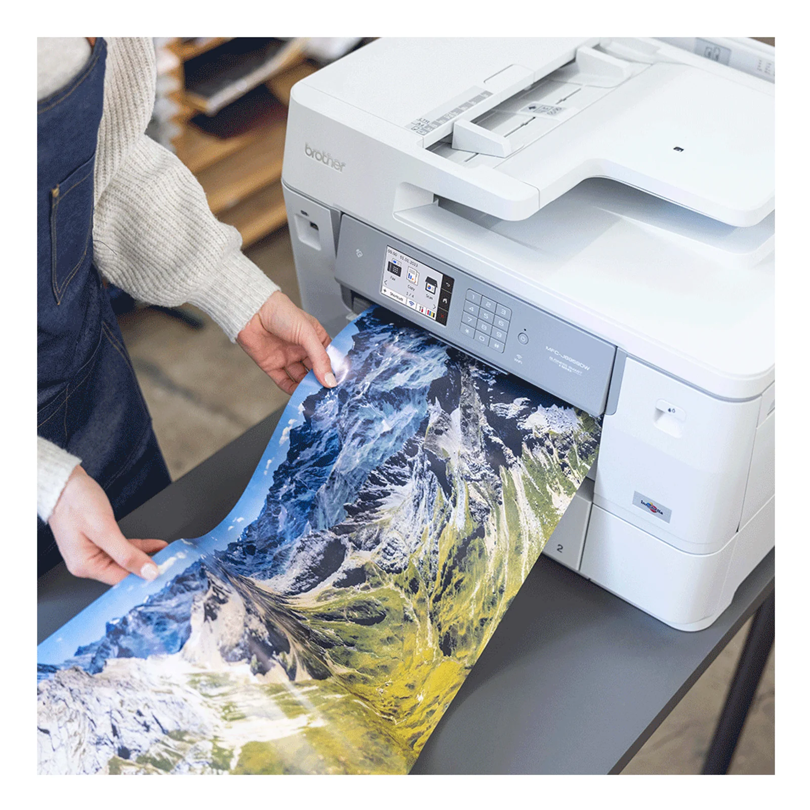 Mains d'utilisateur recuperant un tirage A3 couleur paysage de montagne enneigee sortant du Brother MFC-J6959DW