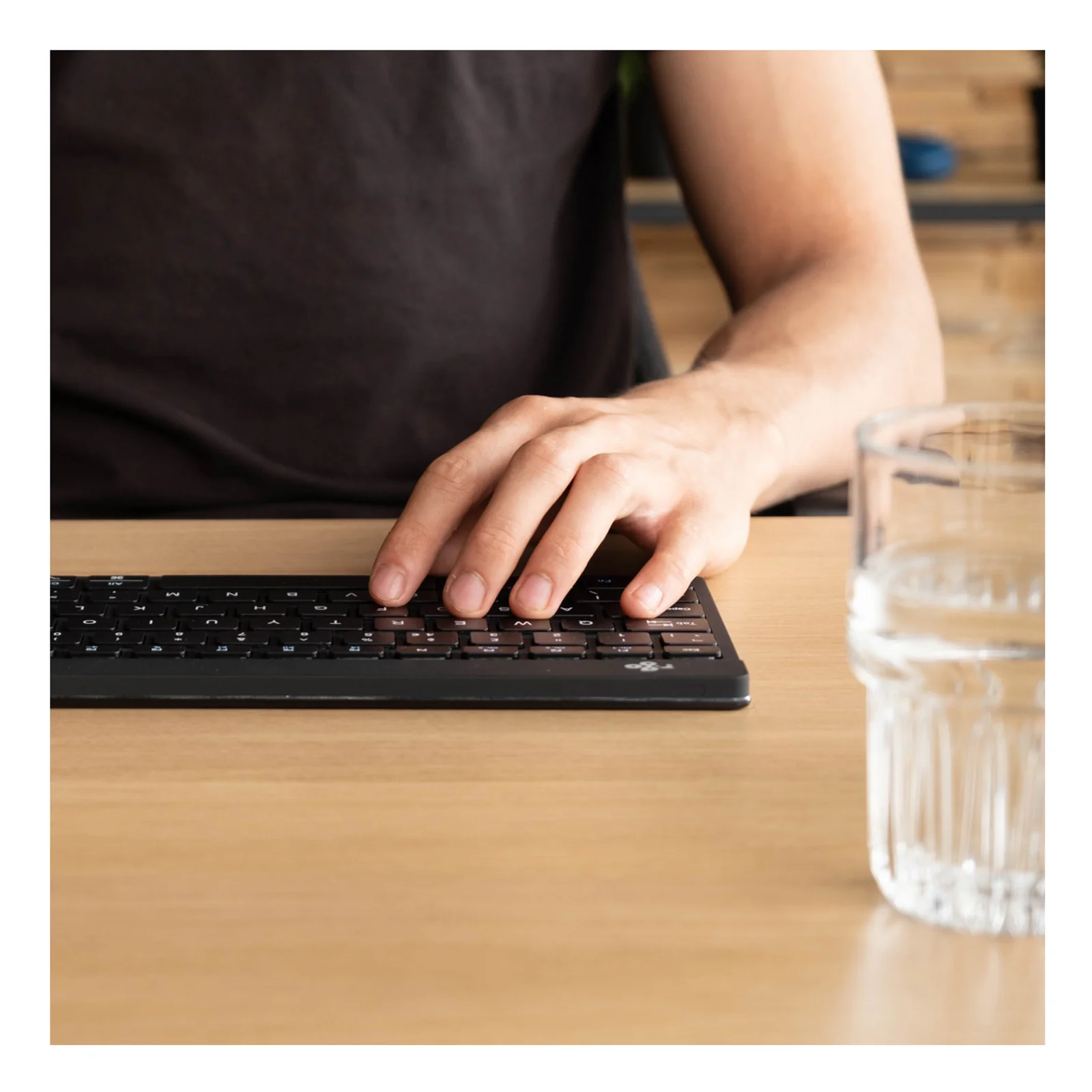 Personne tapant sur le clavier R-Go noir sur bureau bois, verre d'eau posé à droite