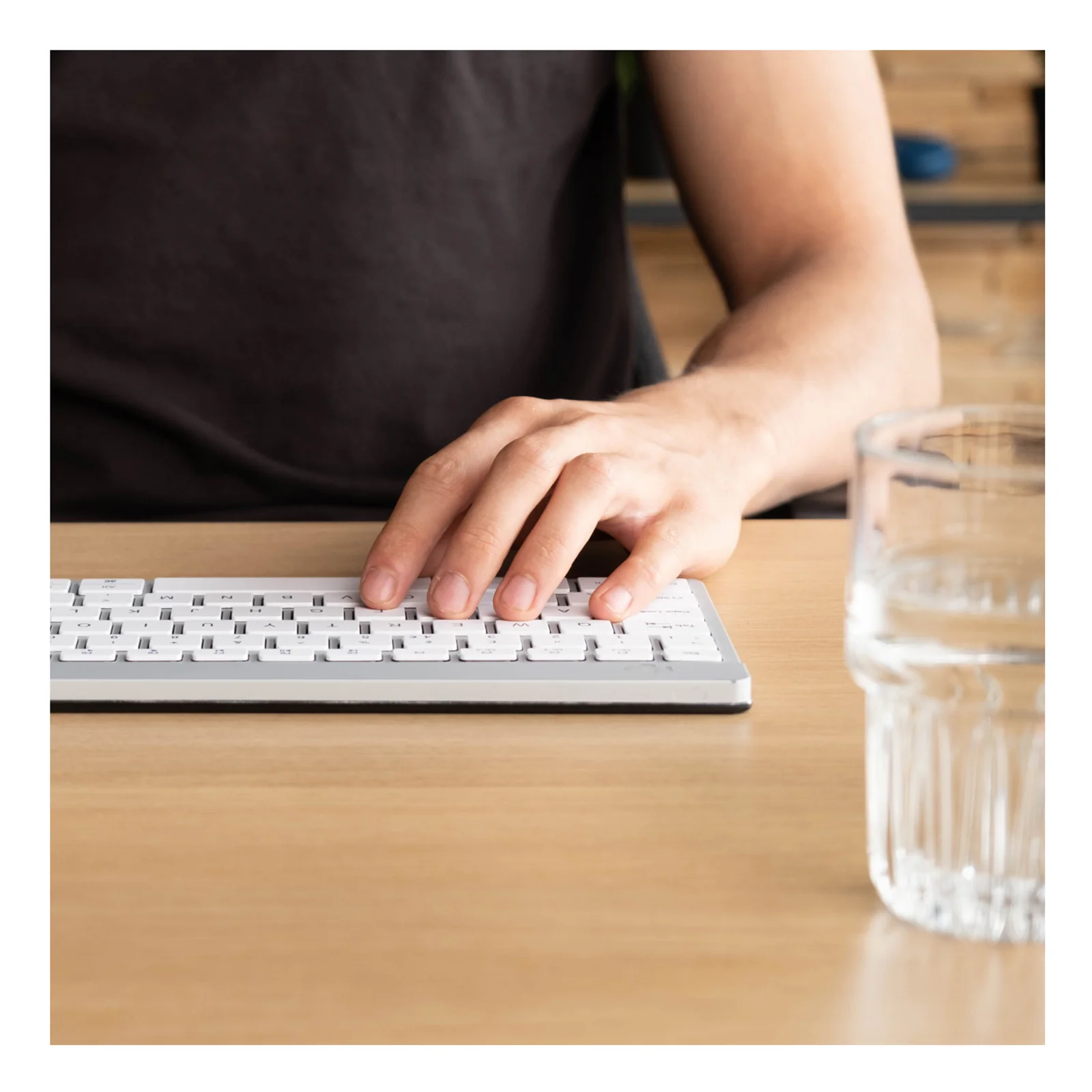 Main en train de taper sur clavier compact blanc, bureau bois, verre d'eau visible à droite