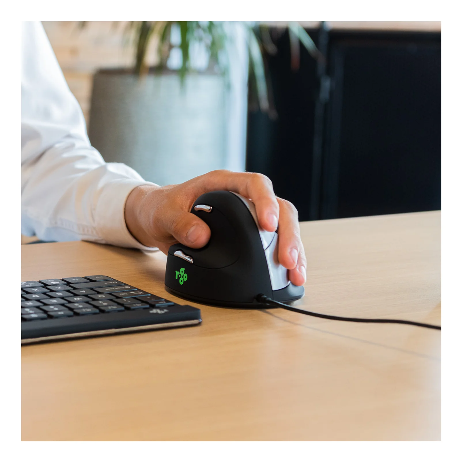 Main gauche tenant la souris verticale R-Go HE Break sur un bureau, clavier et accessoires en arrière-plan