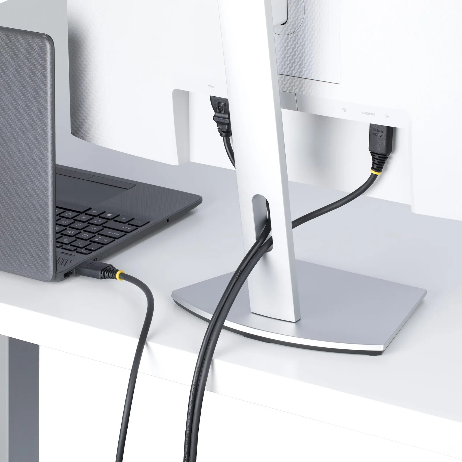 Câble HDMI noir reliant un ordinateur portable à un moniteur blanc posés sur un bureau blanc, deux connecteurs à bague jaune visibles