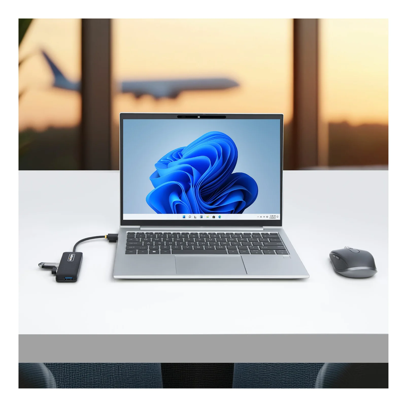 Hub USB-A 4 ports branché sur un ordinateur portable argenté posé sur un bureau, avion visible par la fenêtre en arrière-plan