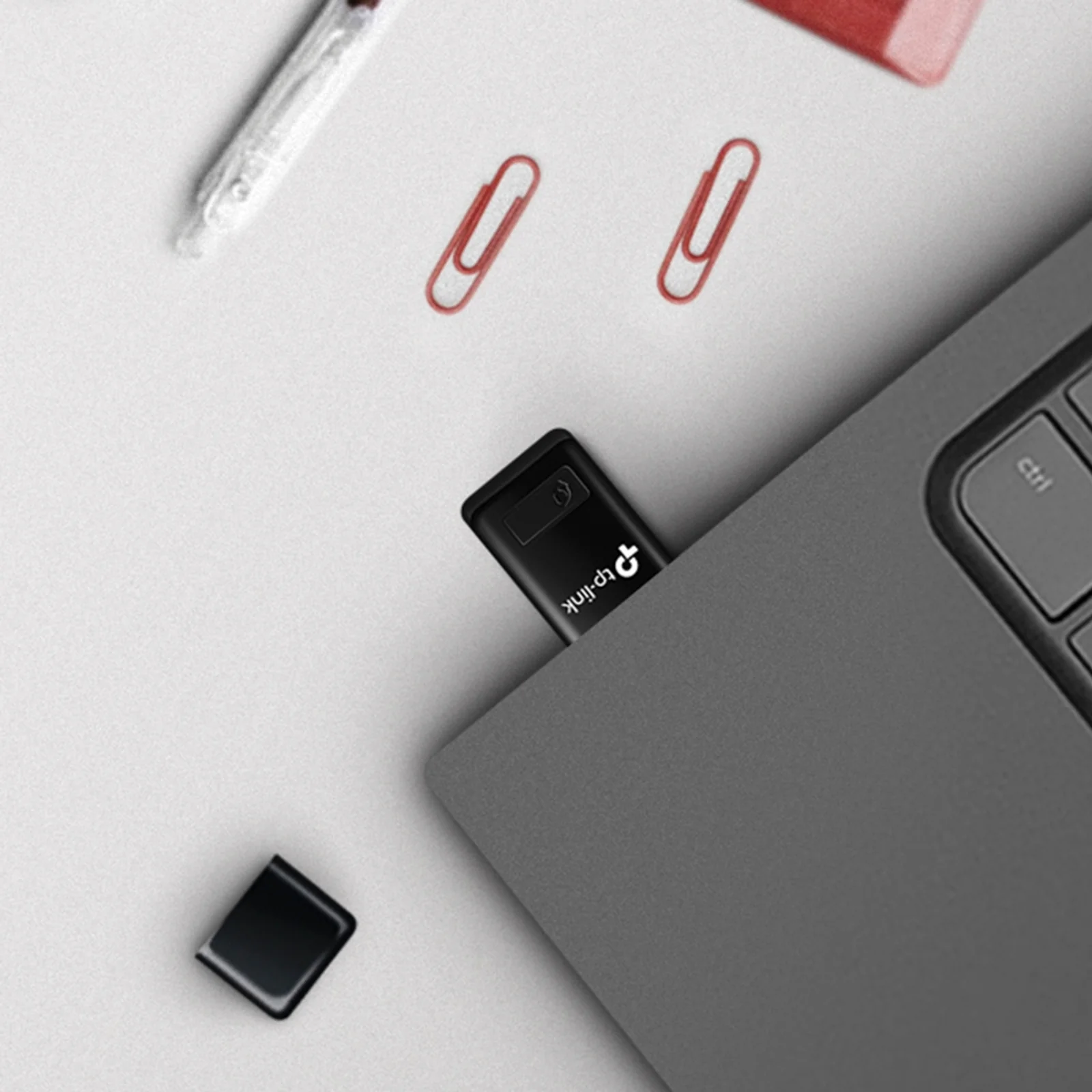 Adaptateur TL-WN823N inséré dans le port USB d'un ordinateur portable gris, capuchon USB posé à côté avec des trombones sur le bureau