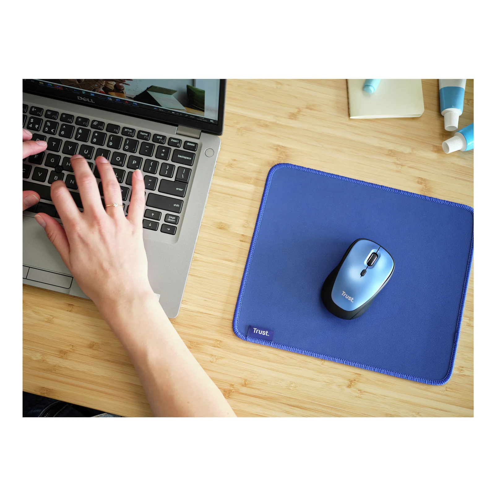 Main posant une souris sur tapis Trust Boye bleu, bureau en bois, laptop visible