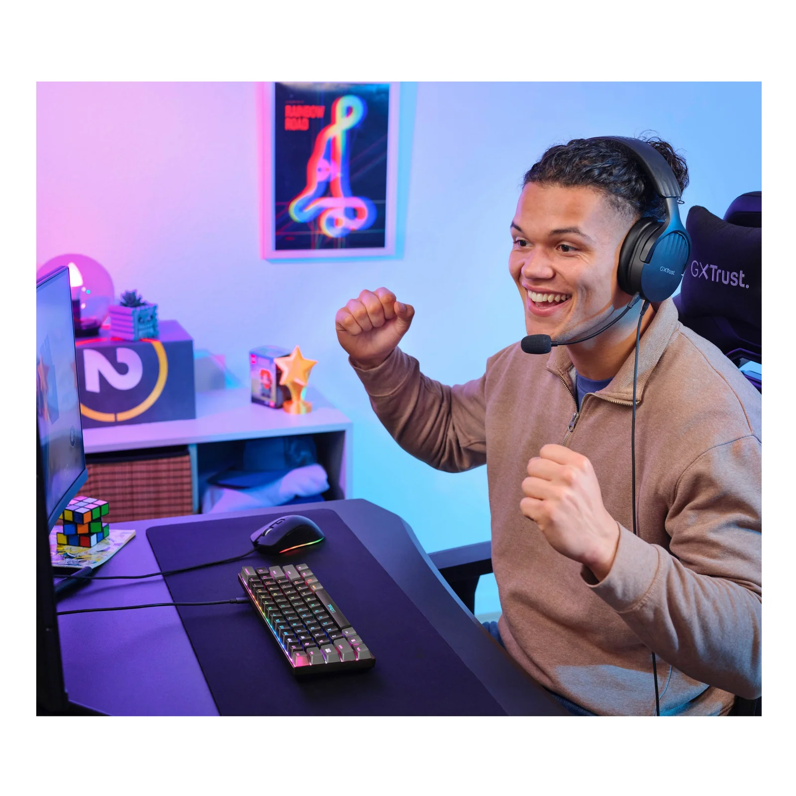 Joueur souriant les bras levés devant son setup gaming, clavier Trust GXT 867 Acira visible sur le bureau