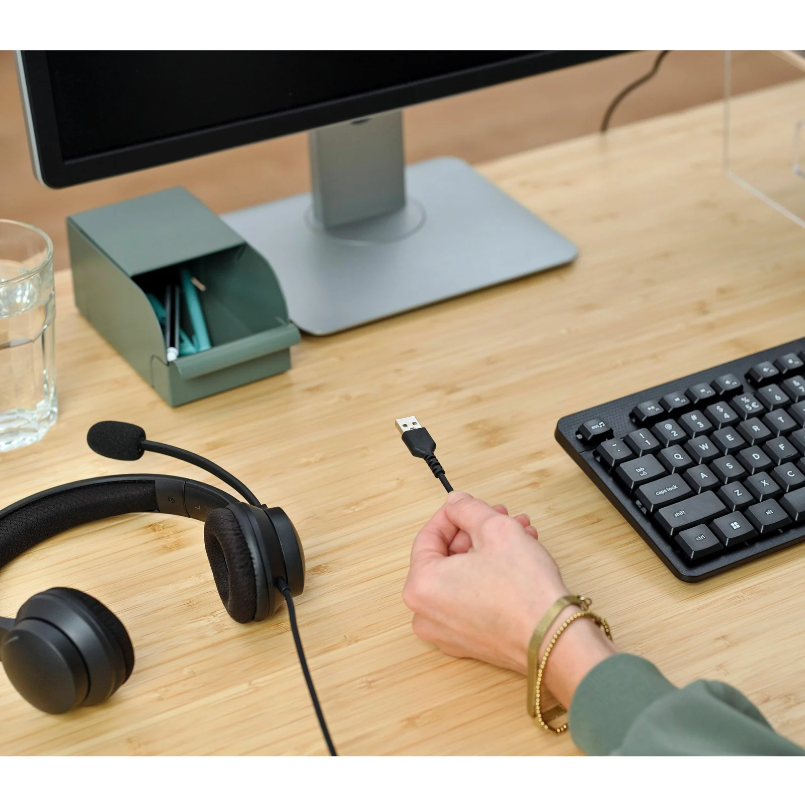 Main tenant le connecteur USB-A du câble Trust HS-260, casque posé près d'un clavier