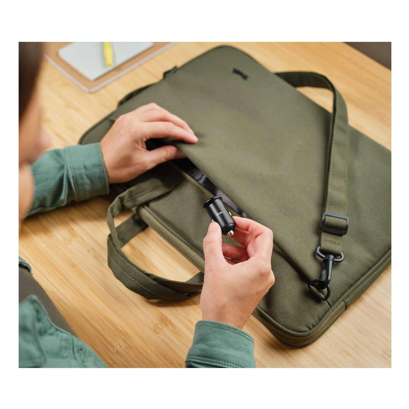 Main tenant le Trust Maxo, chargeur compact noir, avec sac pour ordinateur vert olive en arrière-plan