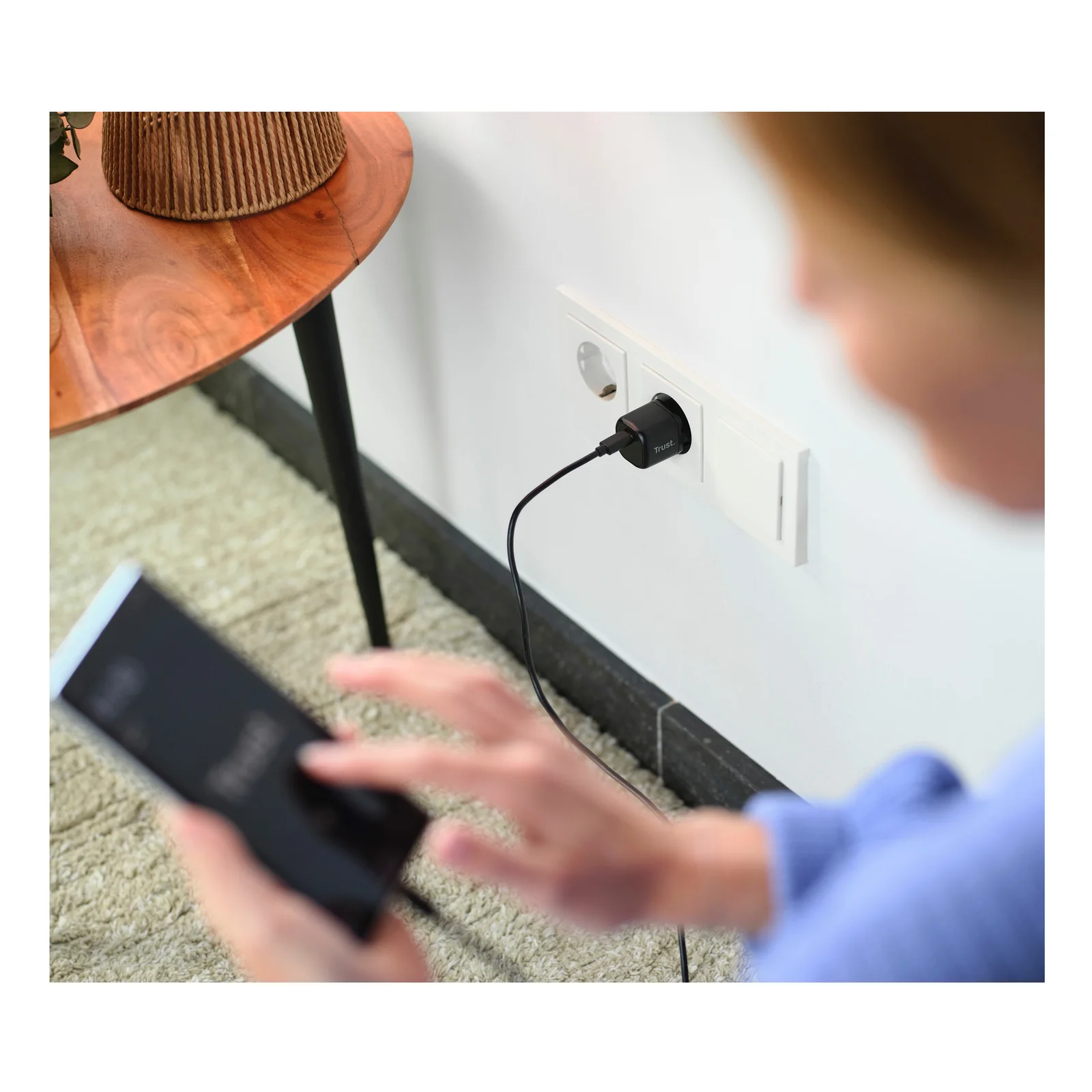 Chargeur noir branché sur une prise murale, personne utilisant un smartphone au second plan