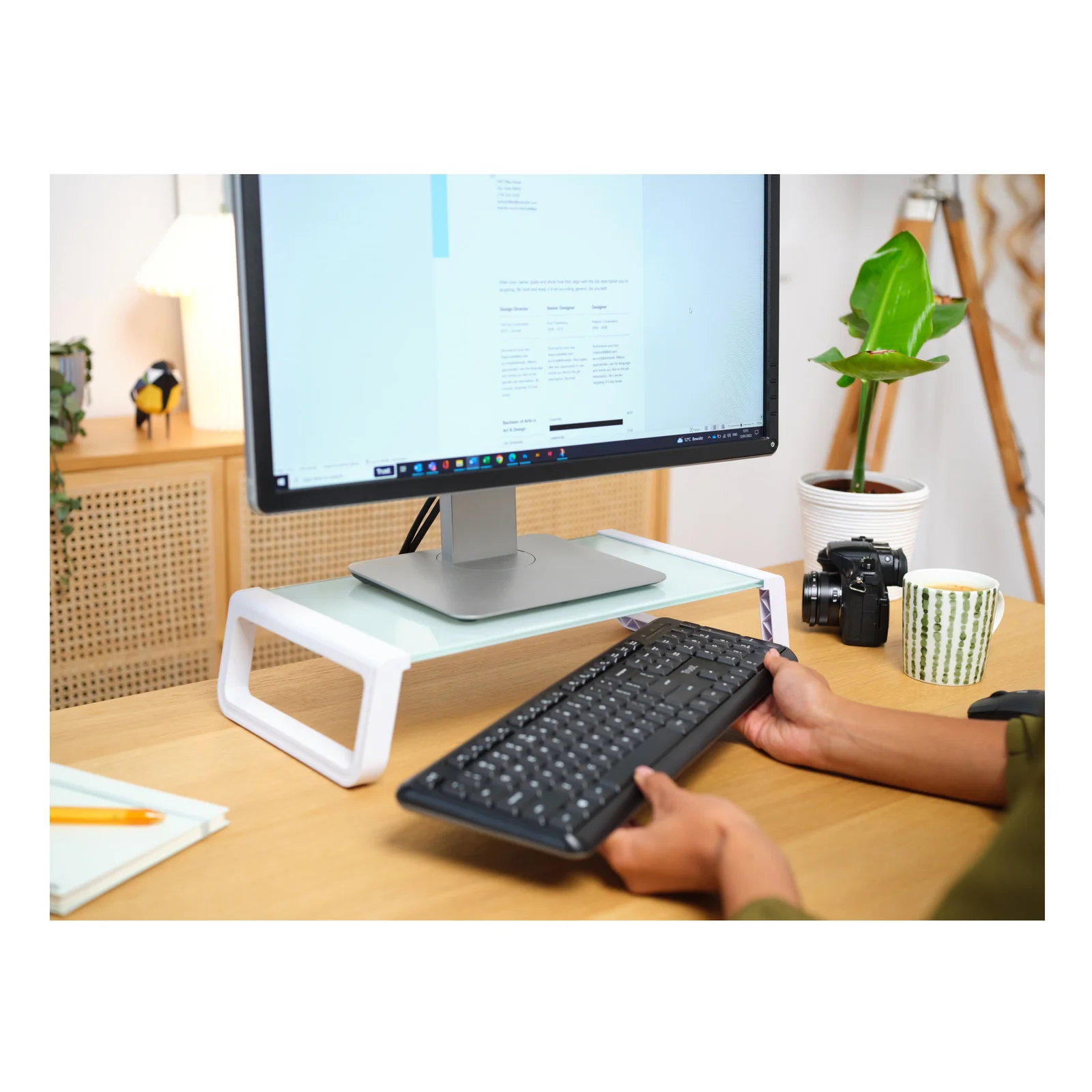 Personne tapant au clavier, moniteur surélevé sur support Trust Monta, plantes en arrière-plan