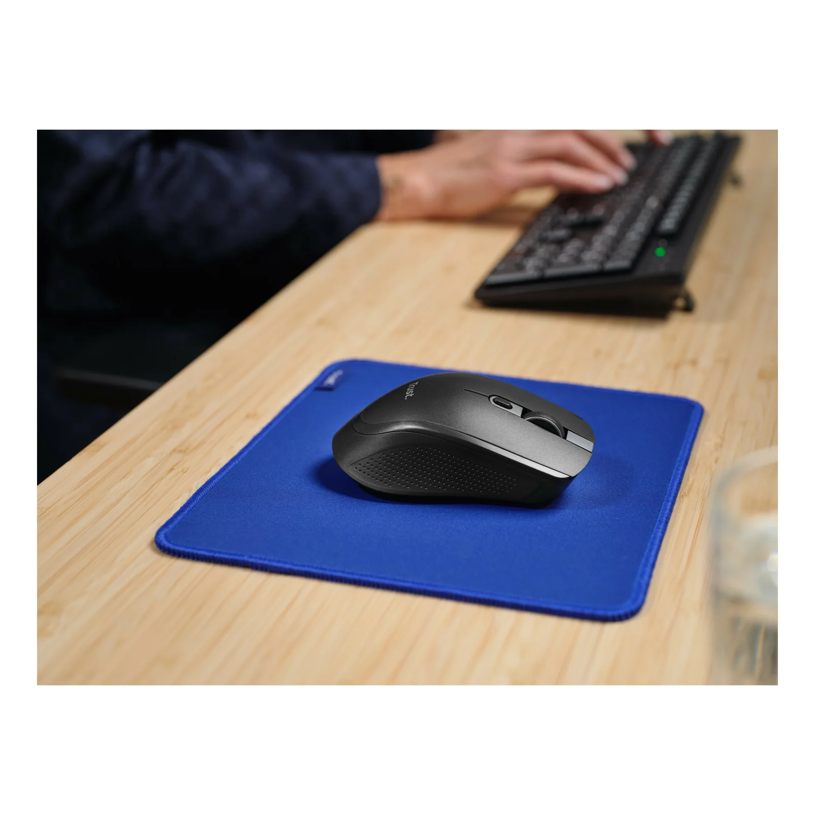 Bureau en bois vu de loin, souris Trust Ozaa+ sur tapis bleu, clavier et verre posés à côté