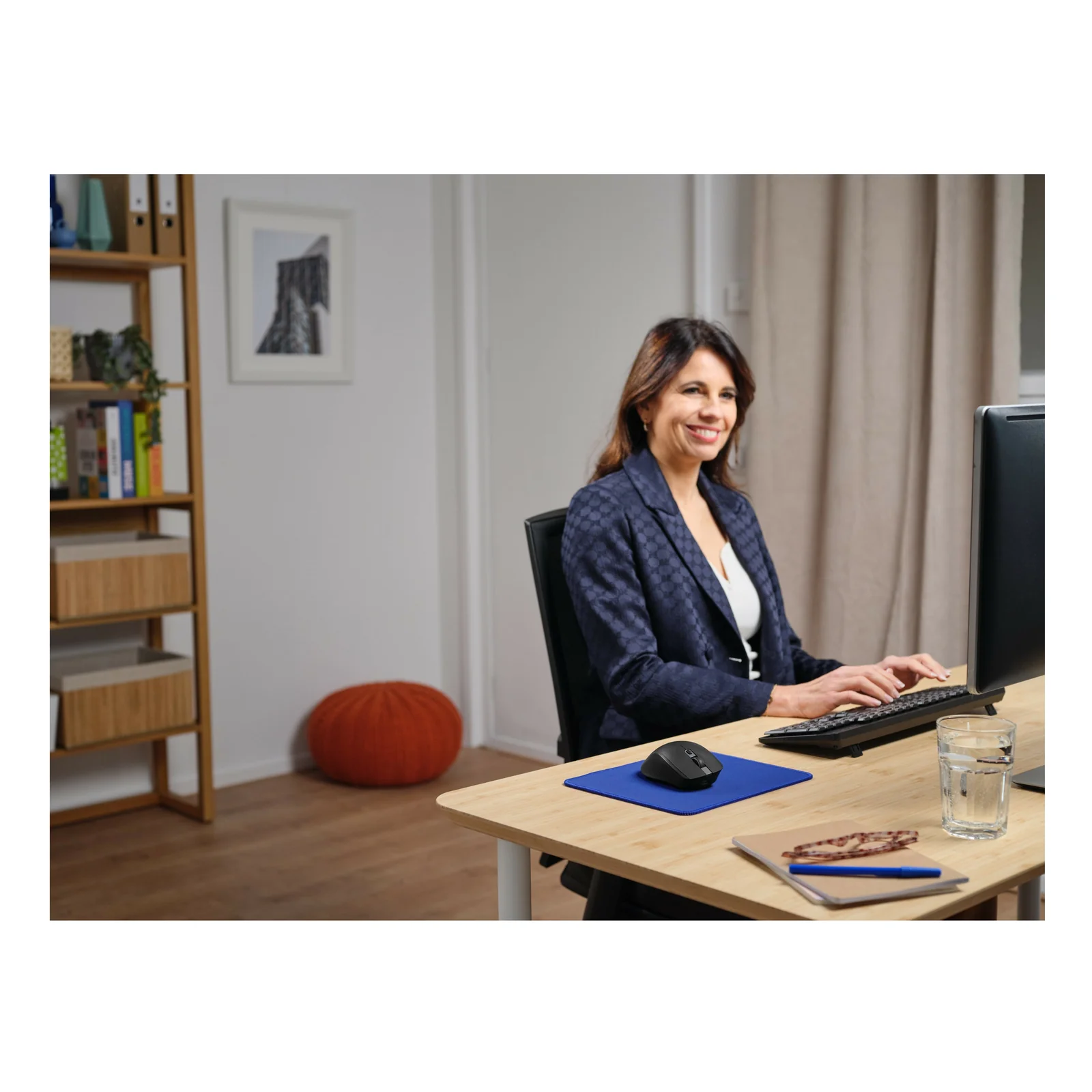 Femme en veste sombre assise à son bureau, souris Trust Ozaa+ sur tapis bleu devant un écran