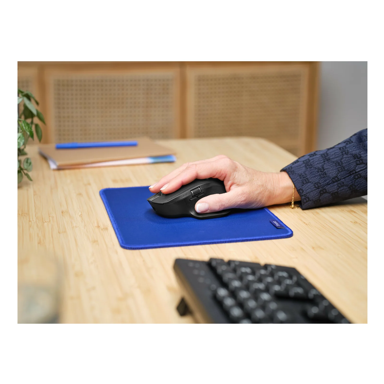 Main posée sur la Trust Ozaa+ noire, tapis de souris bleu sur bureau en bois, clavier au fond