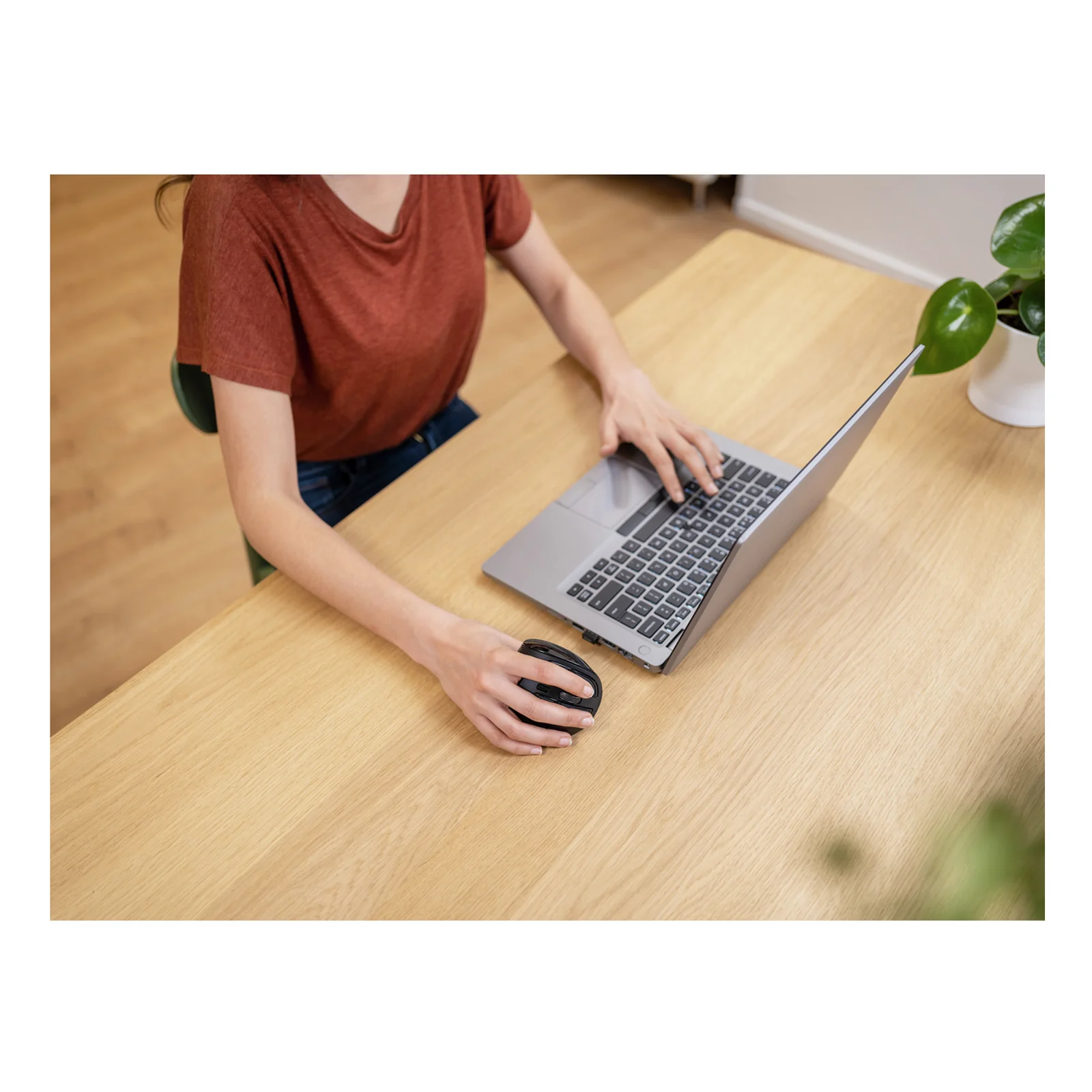 Main droite utilisant la souris Trust Voxx sur un bureau en bois clair, ordinateur portable au fond